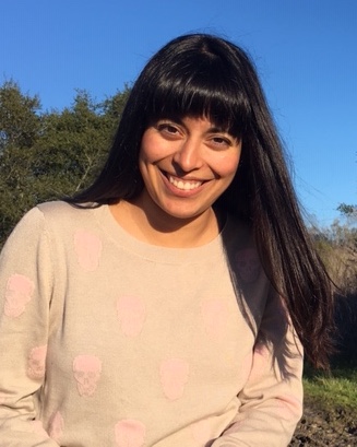 Itzul smiling at the camera. Behind her is a blue sky, field of grass, and many trees. She is wearing a beige long sleeves shirt, and she has her long hair and bangs down.
