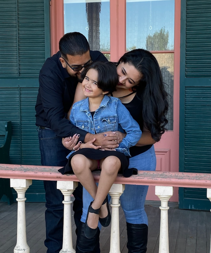 Adriana and her partner hugging their daughter while she sits on a railing, and they stand behind her on a porch. Their daughter is wearing a black dress with a blue jean jacket, while they both wear black.